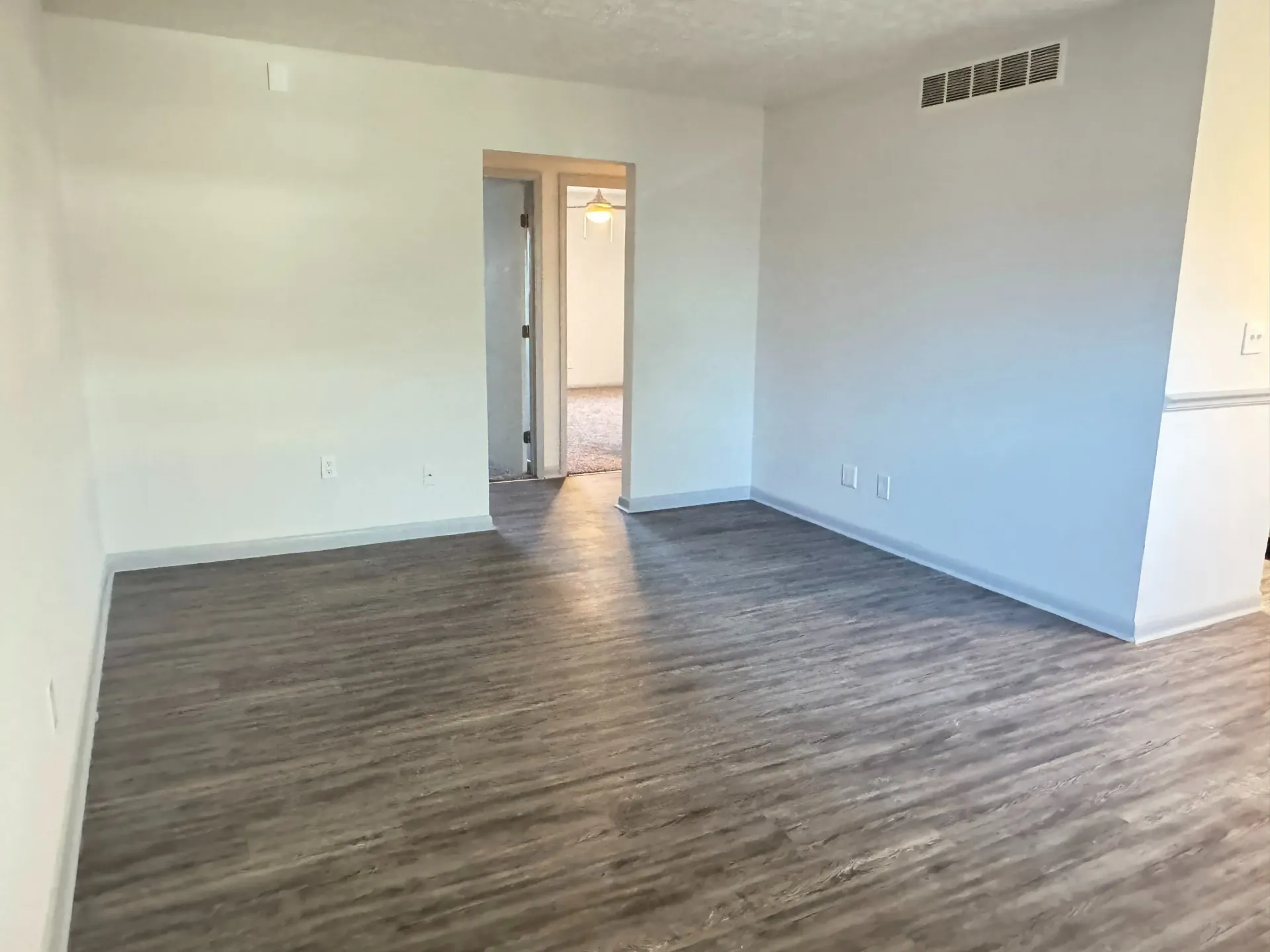 Empty living room with gray wood-look flooring, white walls, and a doorway to a hallway