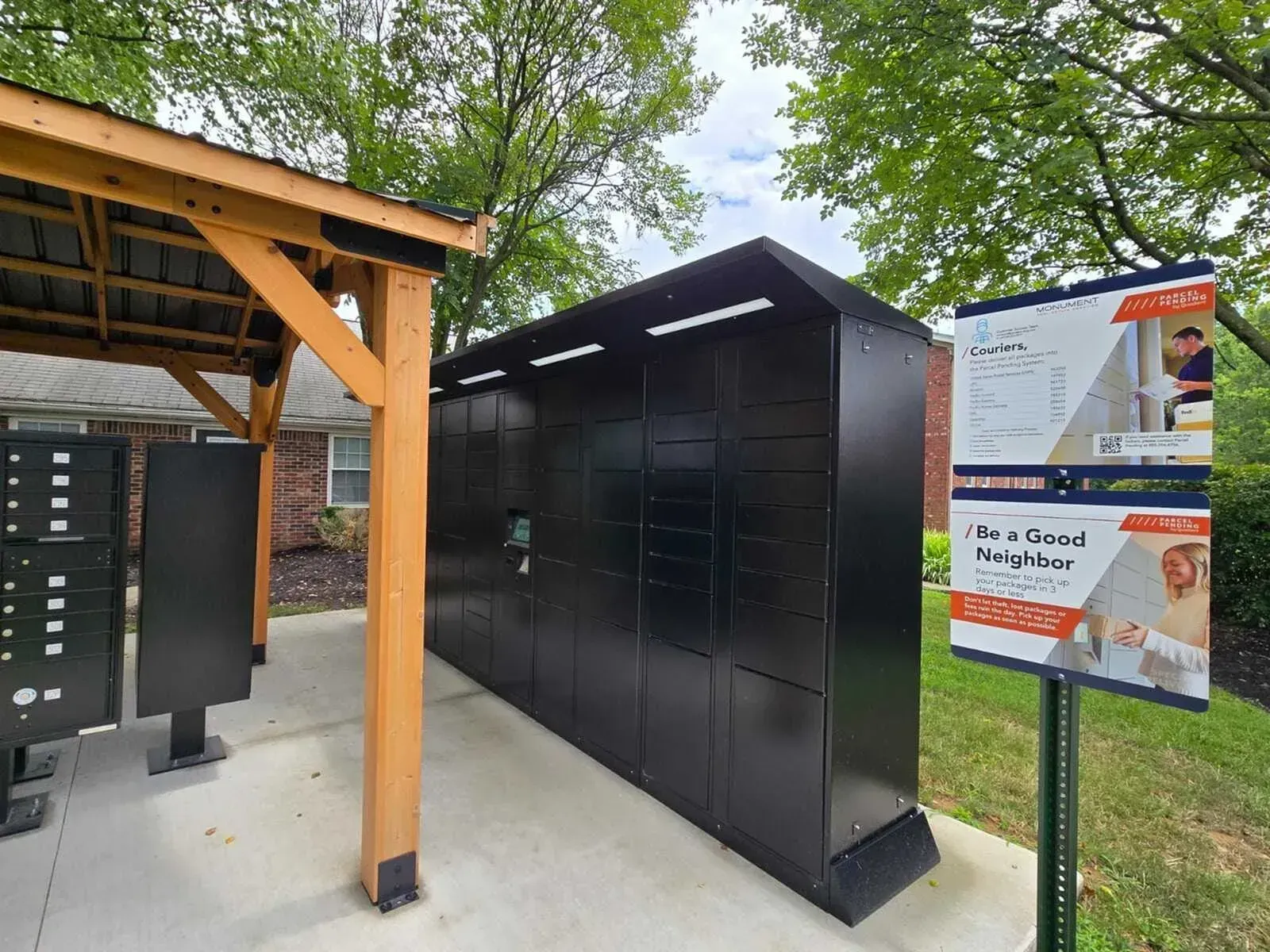 Outdoor black storage lockers beside a covered wooden shelter and a signboard in a park-like setting