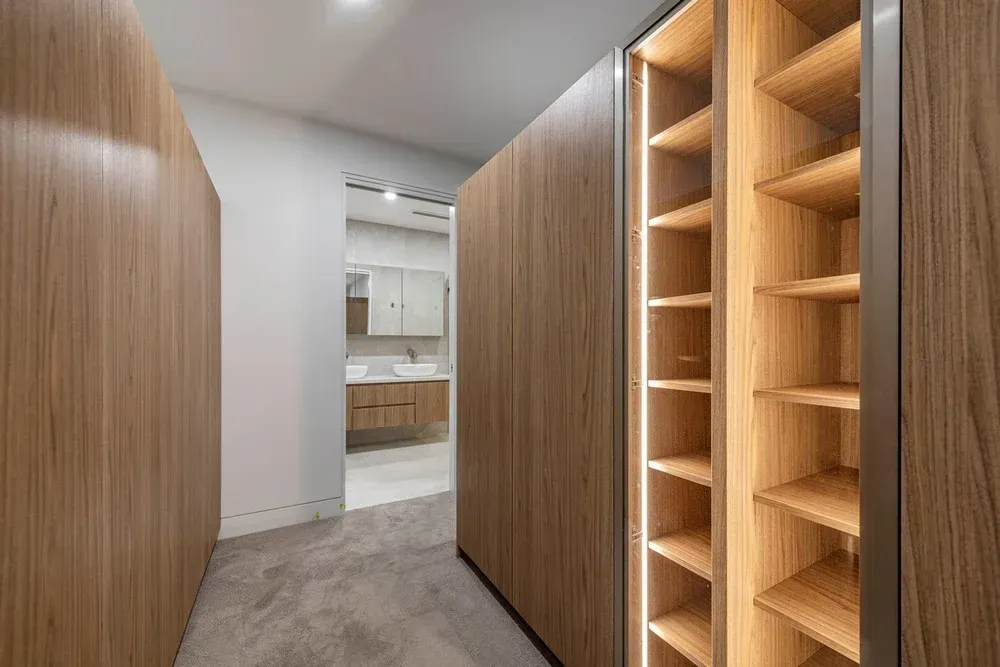 A modern walk-in closet featuring floor-to-ceiling wood cabinetry, illuminated shelves, and a view into a bathroom.