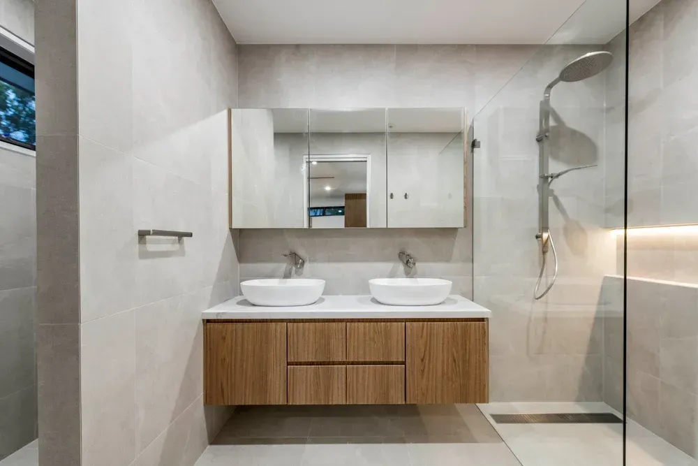 A modern bathroom featuring a floating wooden vanity, two white vessel sinks, a medicine cabinet, and a walk-in shower.