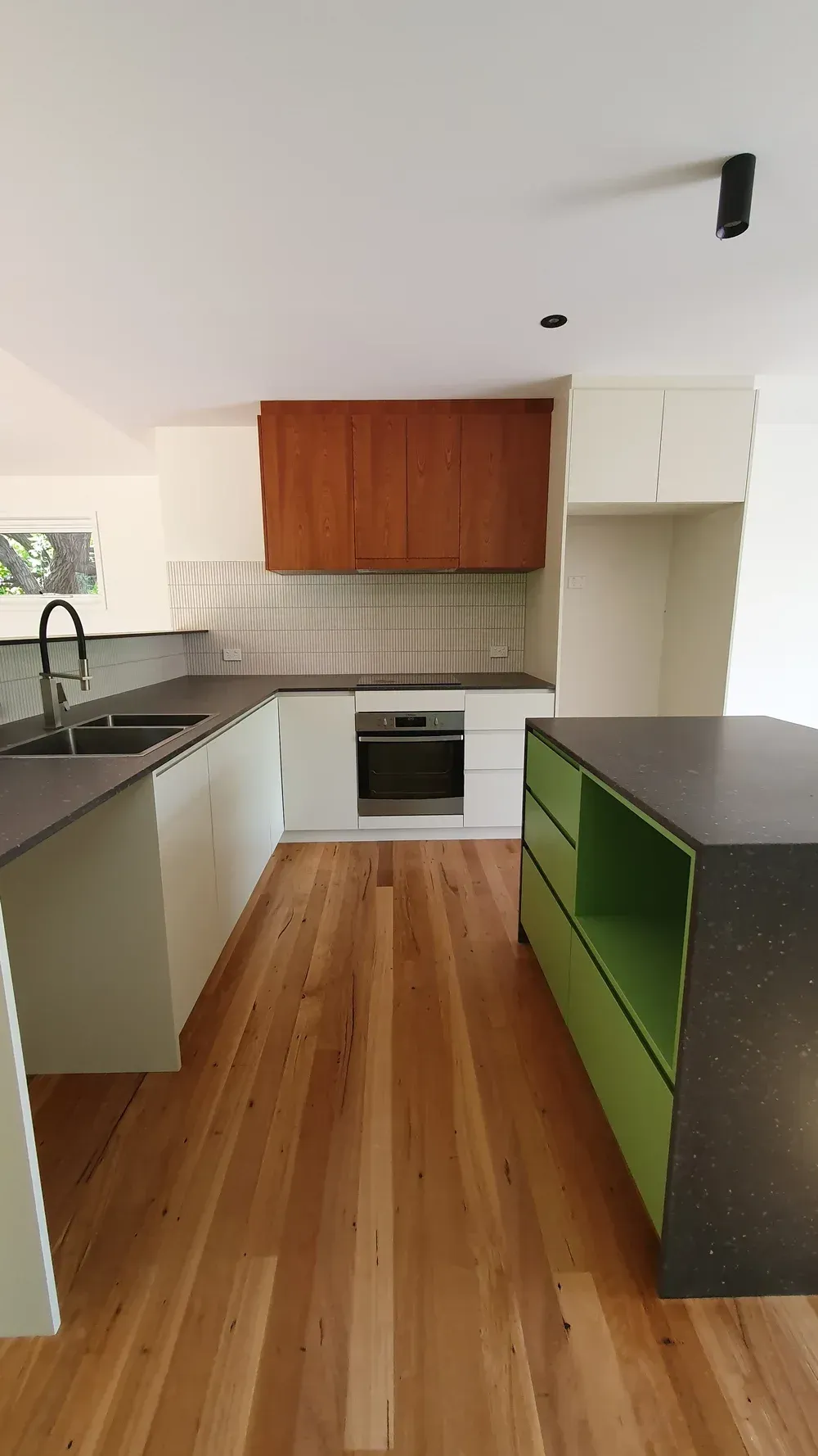 A modern kitchen with a wood-floored galley layout, cream cabinets, a wooden range hood, and a green kitchen island.