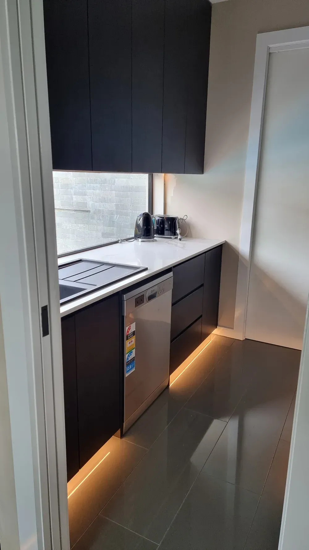 A small, modern kitchenette with dark charcoal cabinets, white countertops, a silver dishwasher, and under-cabinet lighting.
