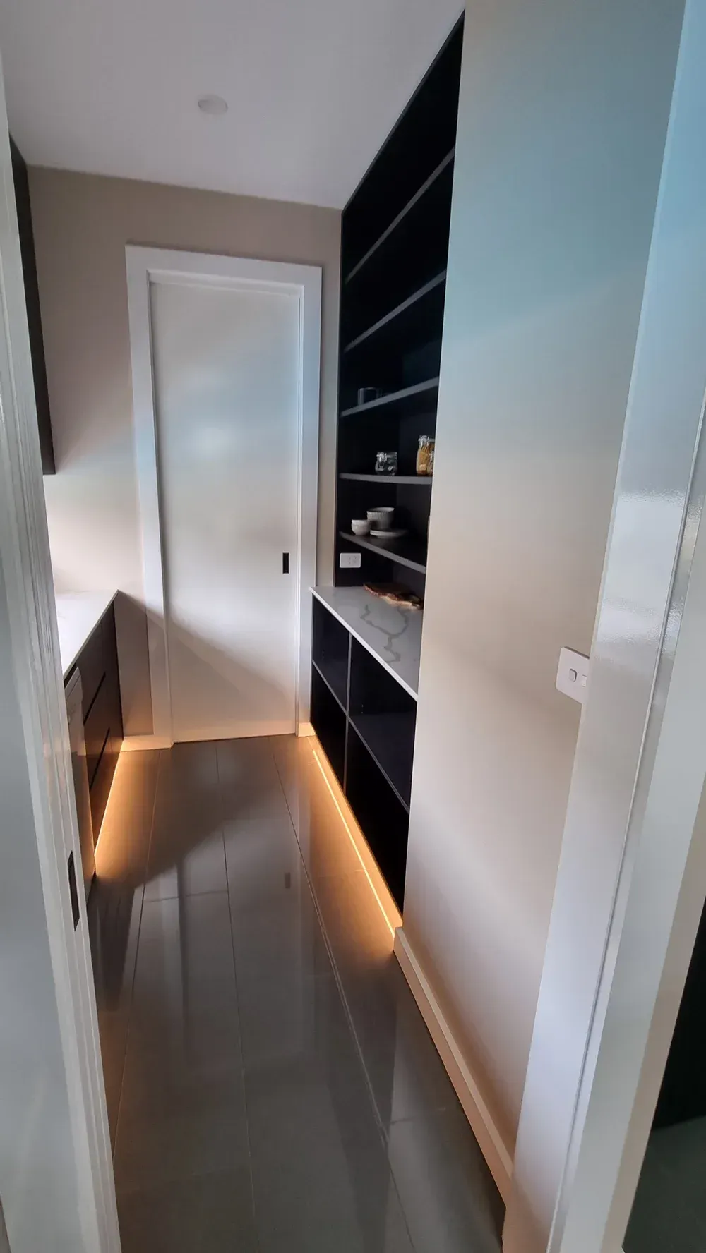 A modern pantry featuring dark shelves, white counters, a white door, and warm floor-level accent lighting.