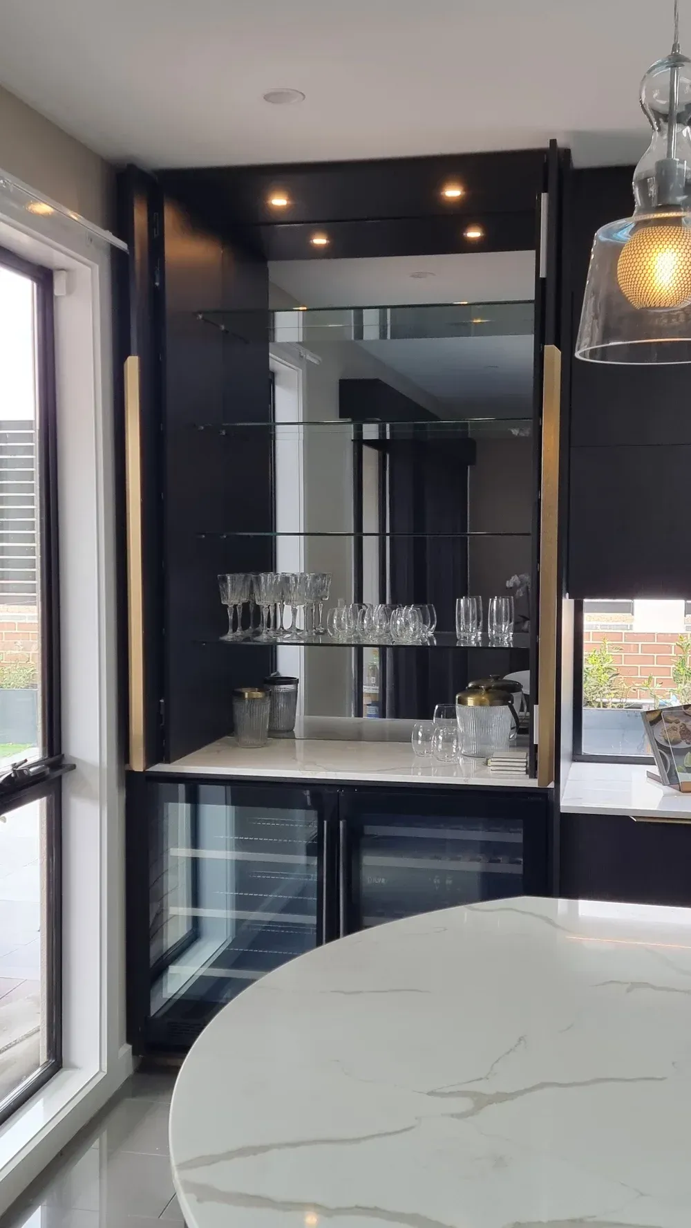 A modern kitchen bar featuring a black cabinet with glass shelving, a mirrored back, built-in wine fridge, and gold accents.