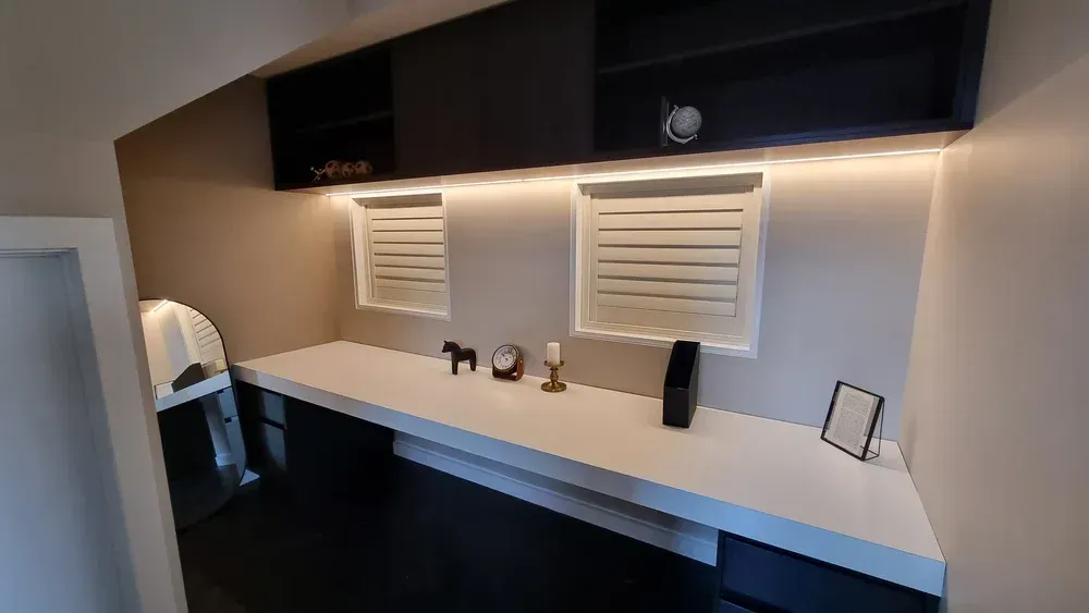 A modern built-in desk area with a white countertop, dark cabinets, two small windows, and soft under-cabinet lighting.