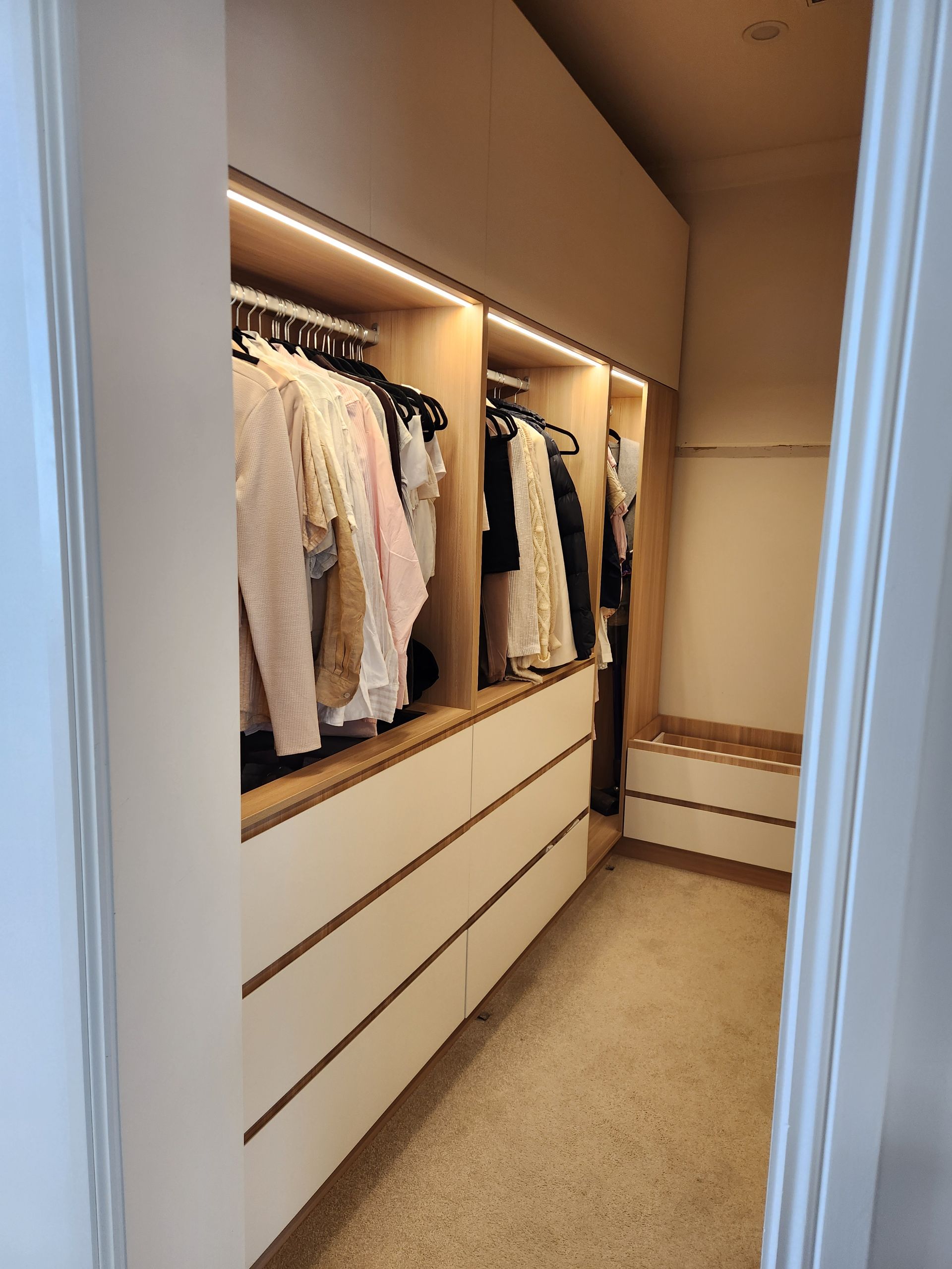 A modern, narrow walk-in pantry with black open shelving, white countertop, and warm LED floor lighting.