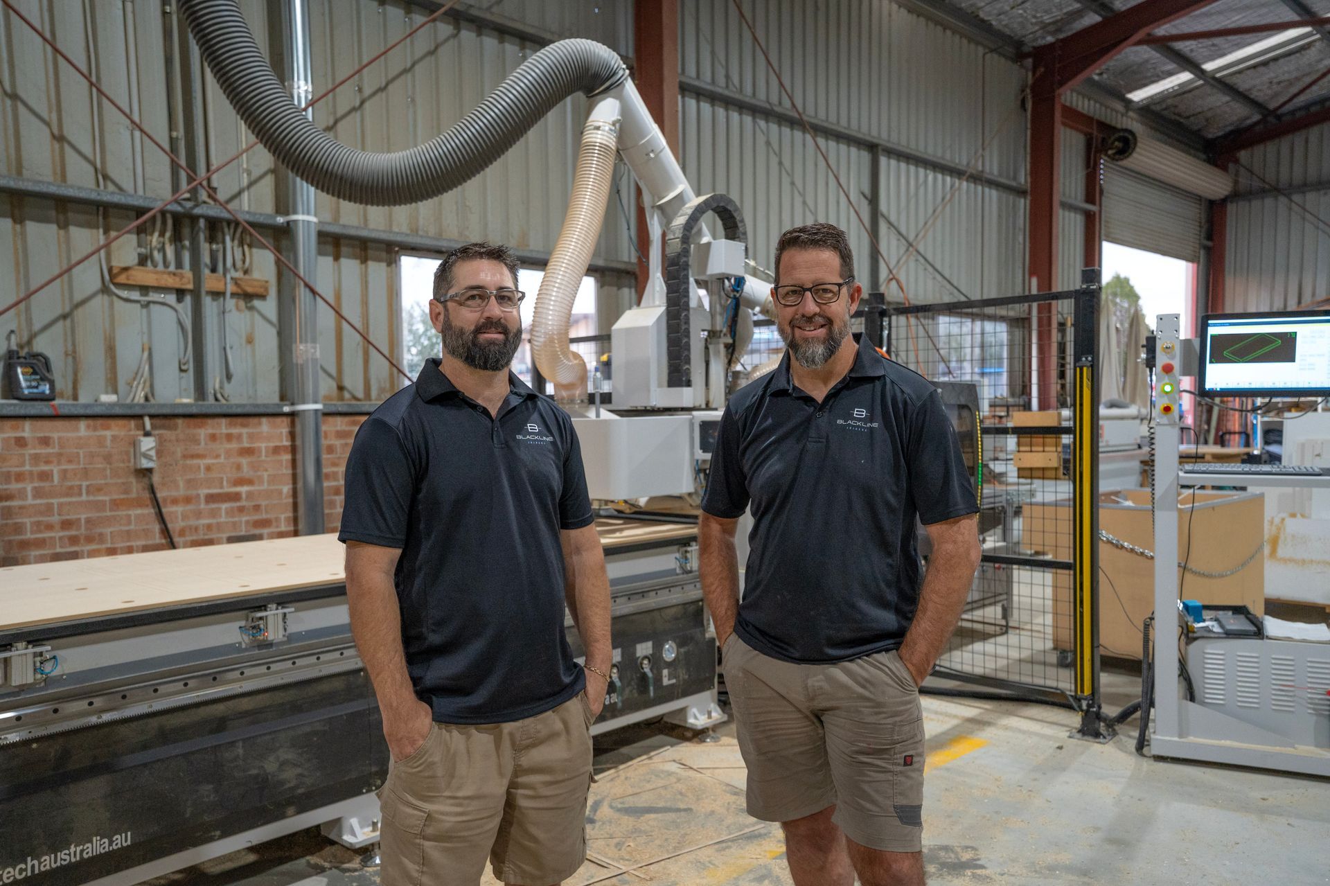 Two people standing in a manufacturing workshop next to a large industrial CNC machine with a vacuum hose attached.