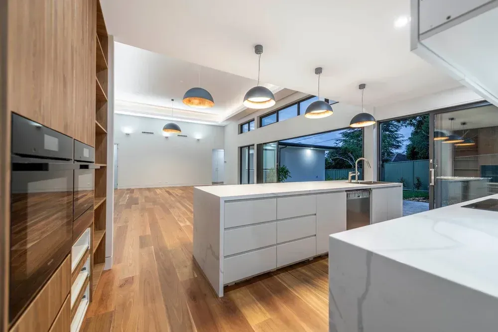 Modern white kitchen with wood floors, island, built-in oven, and large windows overlooking a patio