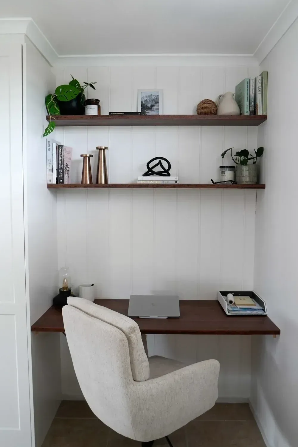 Small home office nook with white chair, wooden desk, shelves, plants, and a laptop in a white alcove