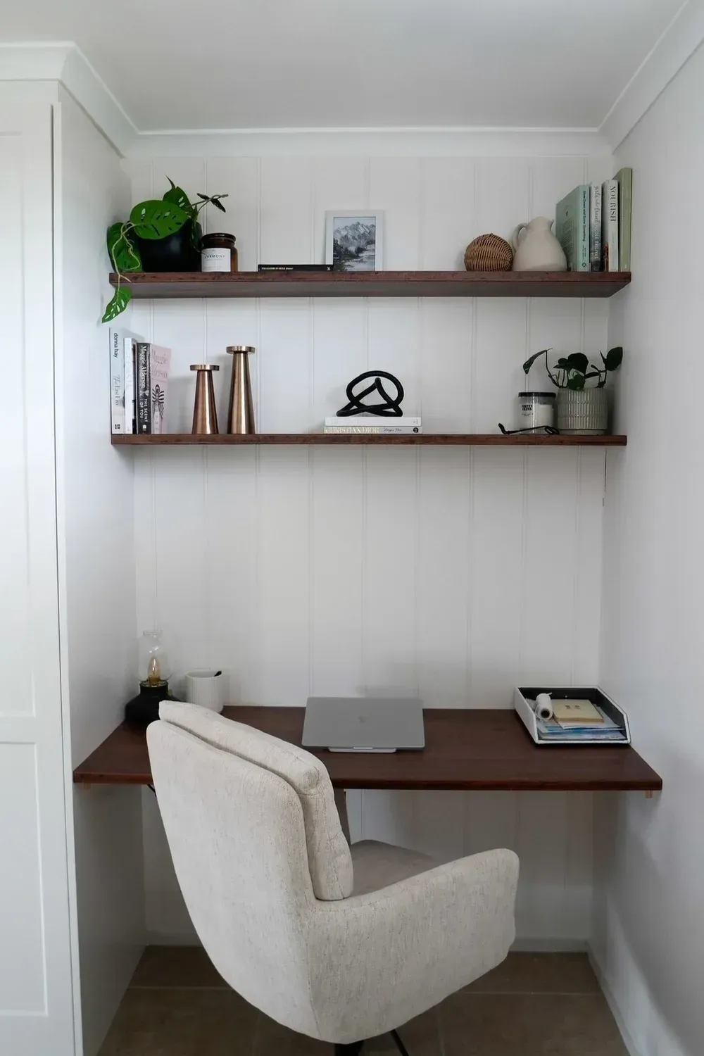 A home office nook with a dark wood desk and two matching shelves against white shiplap walls, featuring a cream chair.