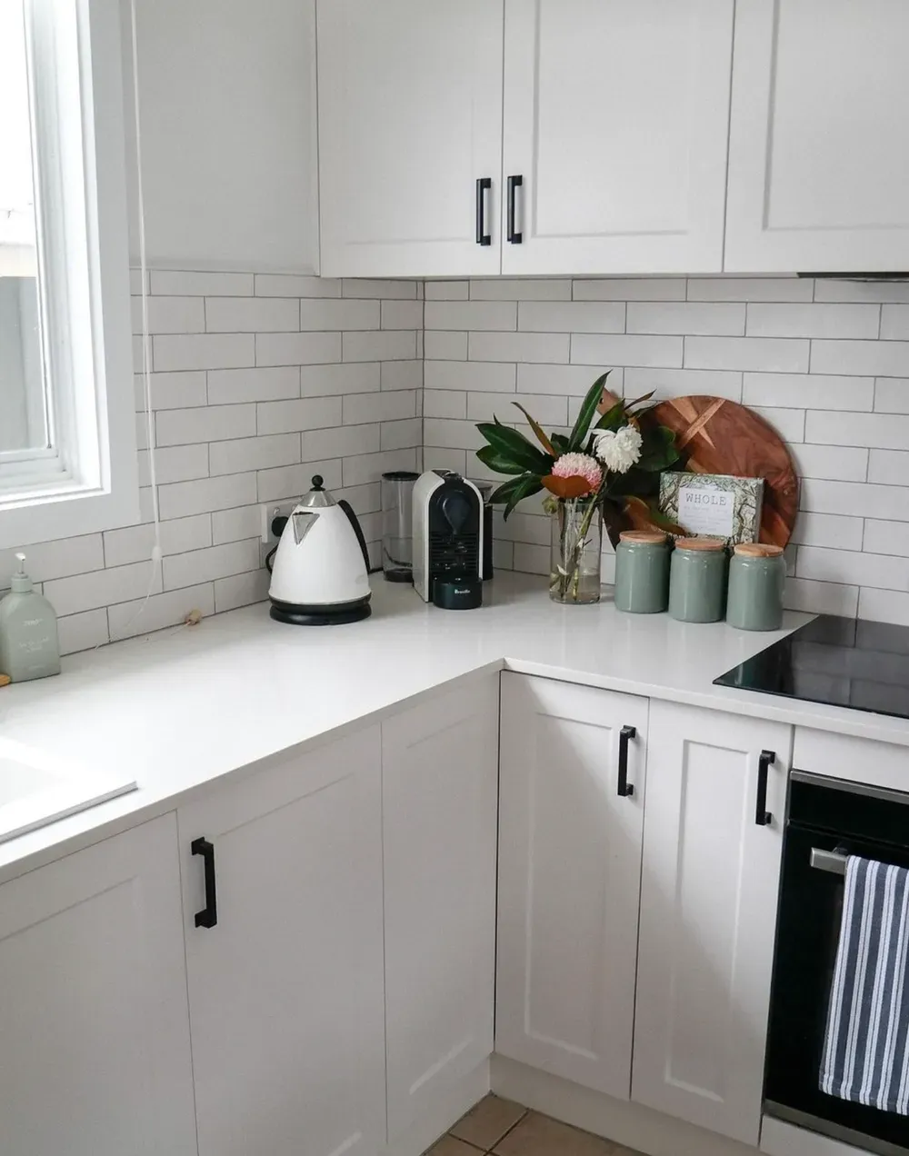 A clean, white modern kitchen with white subway tile backsplash, white cabinets with black handles, and a coffee station.