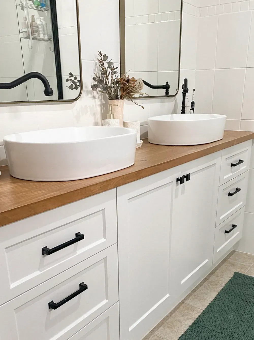 A modern bathroom vanity with a wood countertop, white cabinets, black hardware, two vessel sinks, and arched mirrors.
