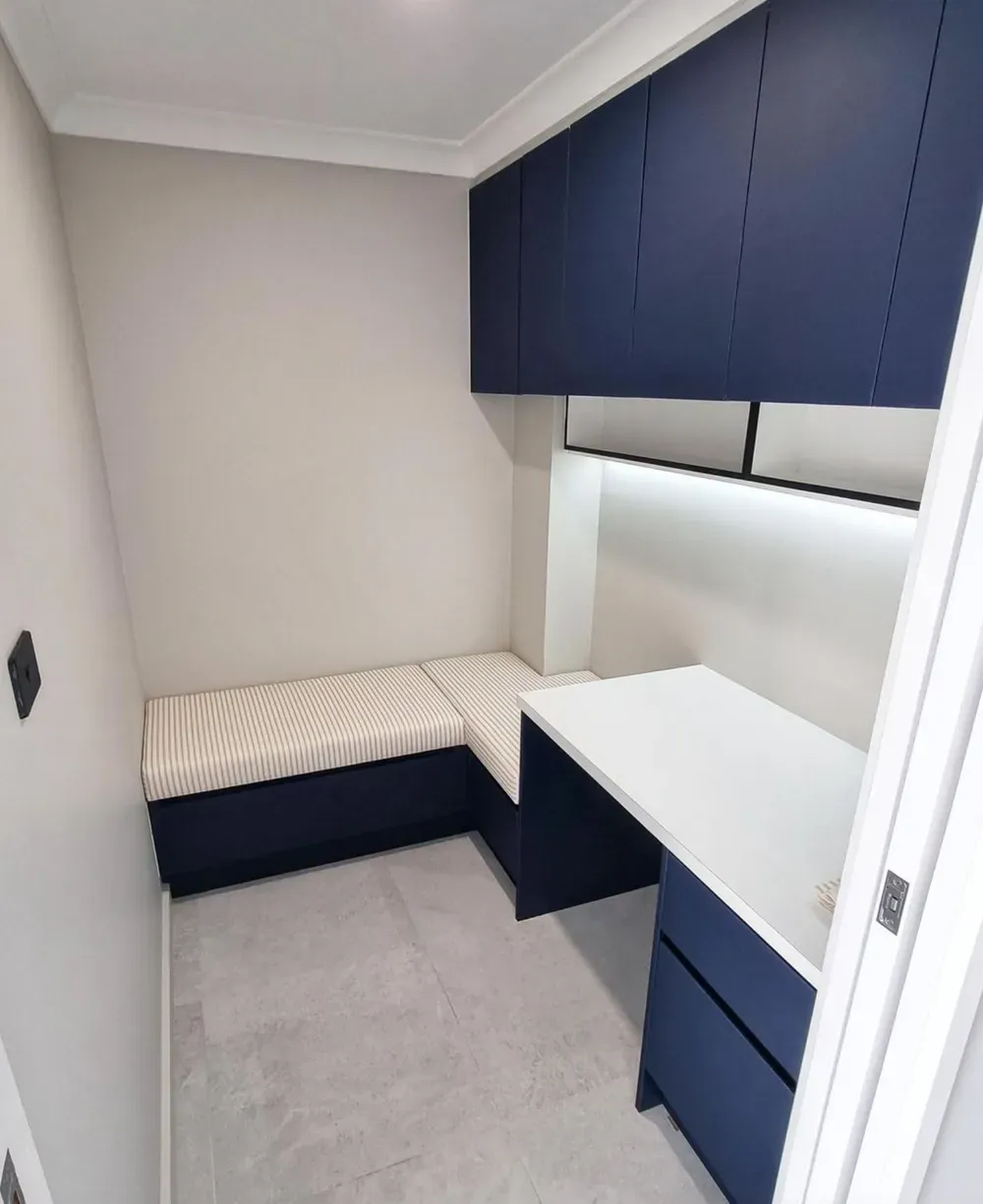 A small home office nook with dark blue cabinets, a white desk, and a cushioned bench seat against light gray walls.