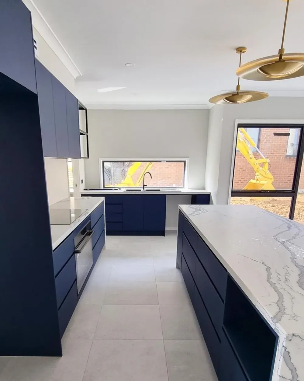 A modern kitchen with dark blue cabinetry, a white marble-topped island, light stone floors, and gold pendant lights.
