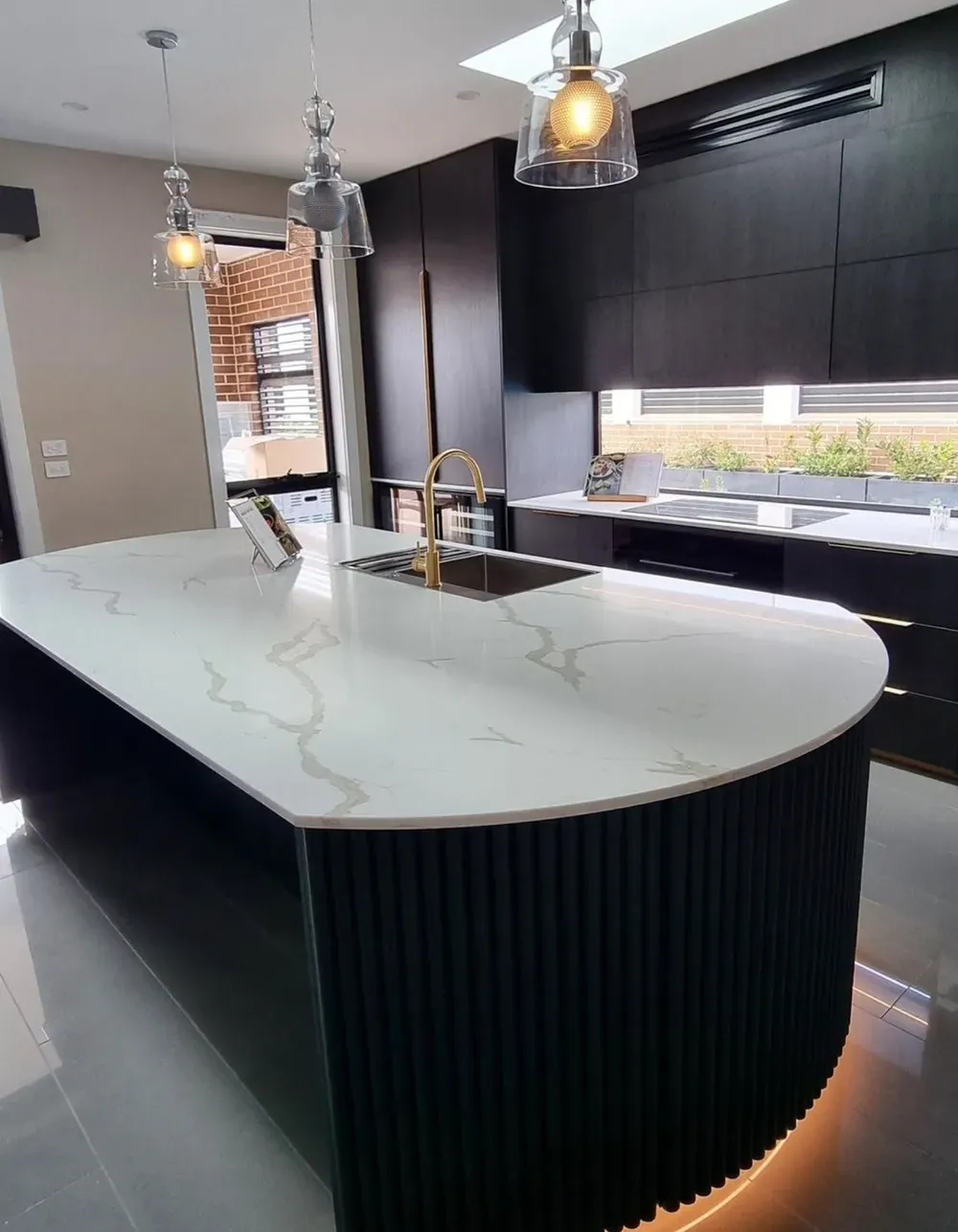 A modern kitchen island with white veined stone countertops and a dark, fluted curved base, featuring gold fixtures.
