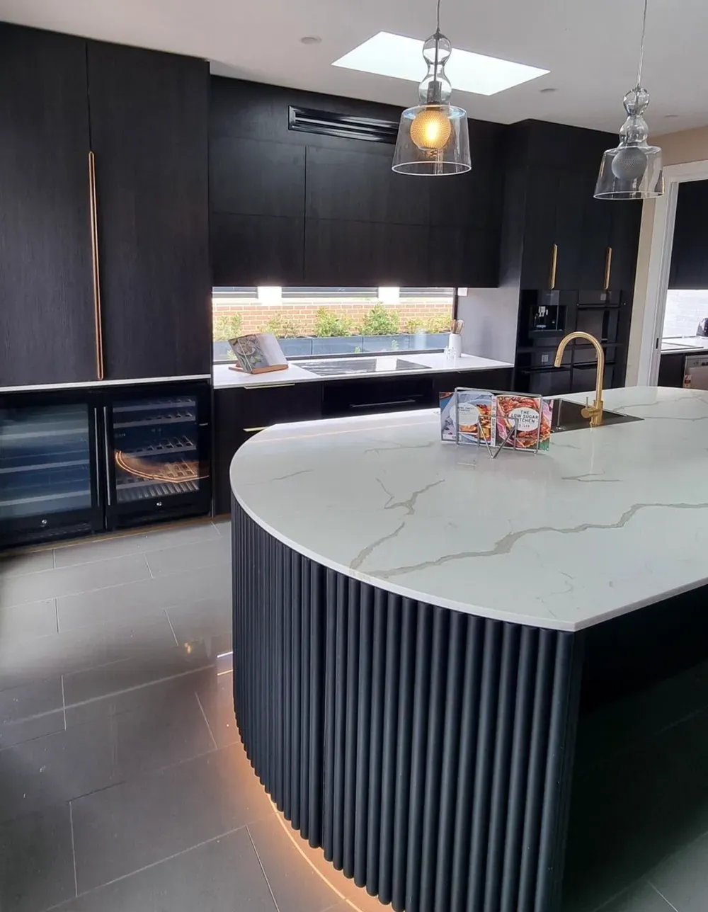 A modern kitchen featuring a curved island with black slatted base and white marble countertop, dark cabinetry, and lighting.