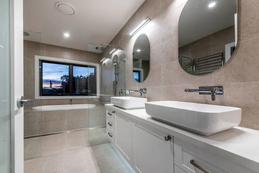 A modern, neutral-toned bathroom with dual white vessel sinks, a glass-enclosed shower, and a large soaking tub by a window.