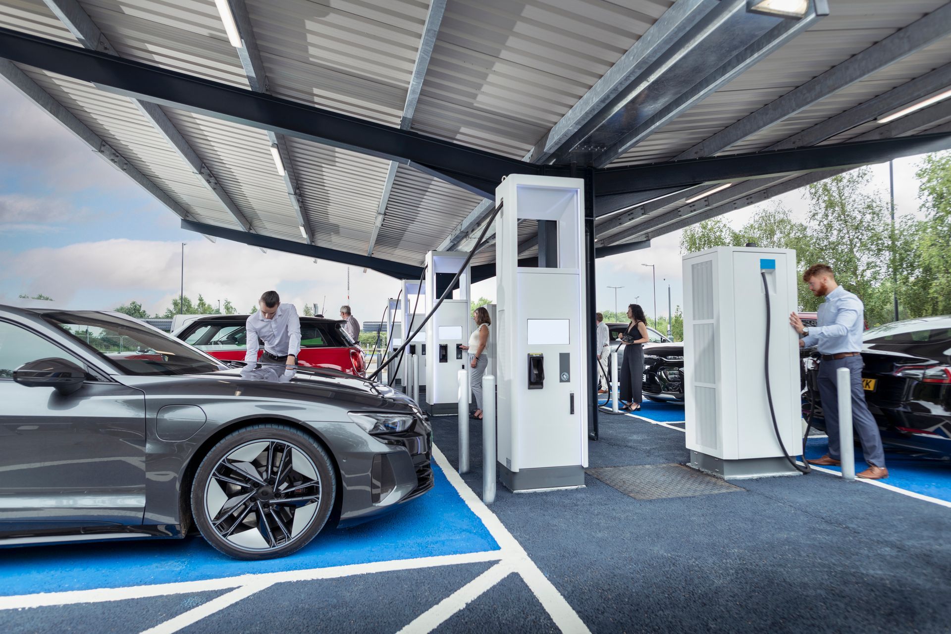 Electric vehicle charging station with cars, people plugging in, under a covered structure.