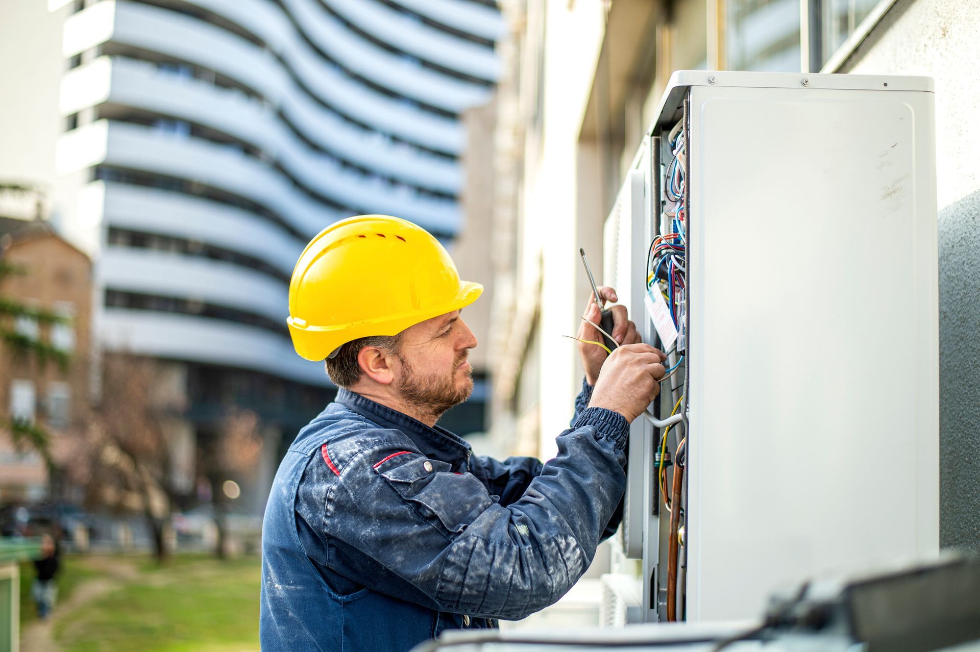 Electrician in yellow hard hat, working on a panel outdoors. Building background.