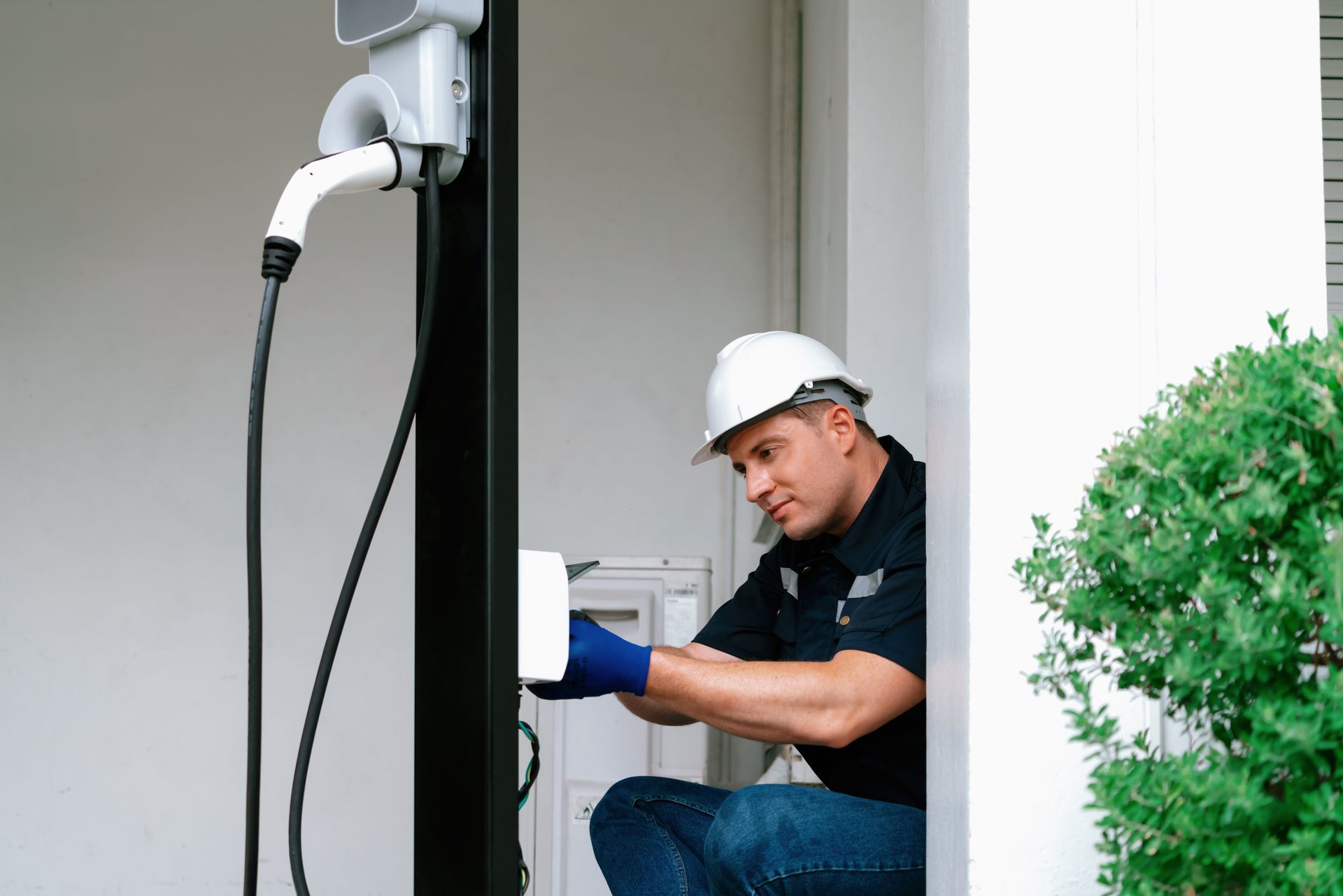 Electrician installing EV charging station on a black pole; white hard hat, blue gloves, neutral expression.