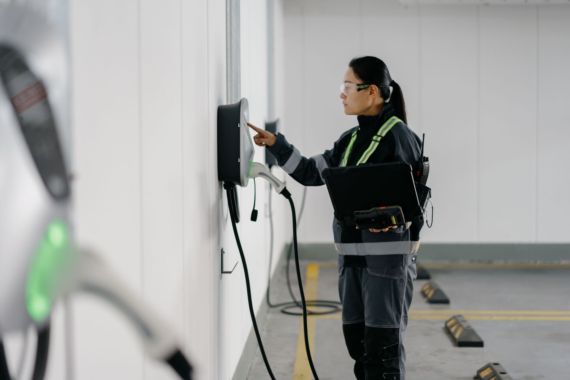 Technician checking EV charging station. Wearing safety vest and holding a laptop in a parking garage.