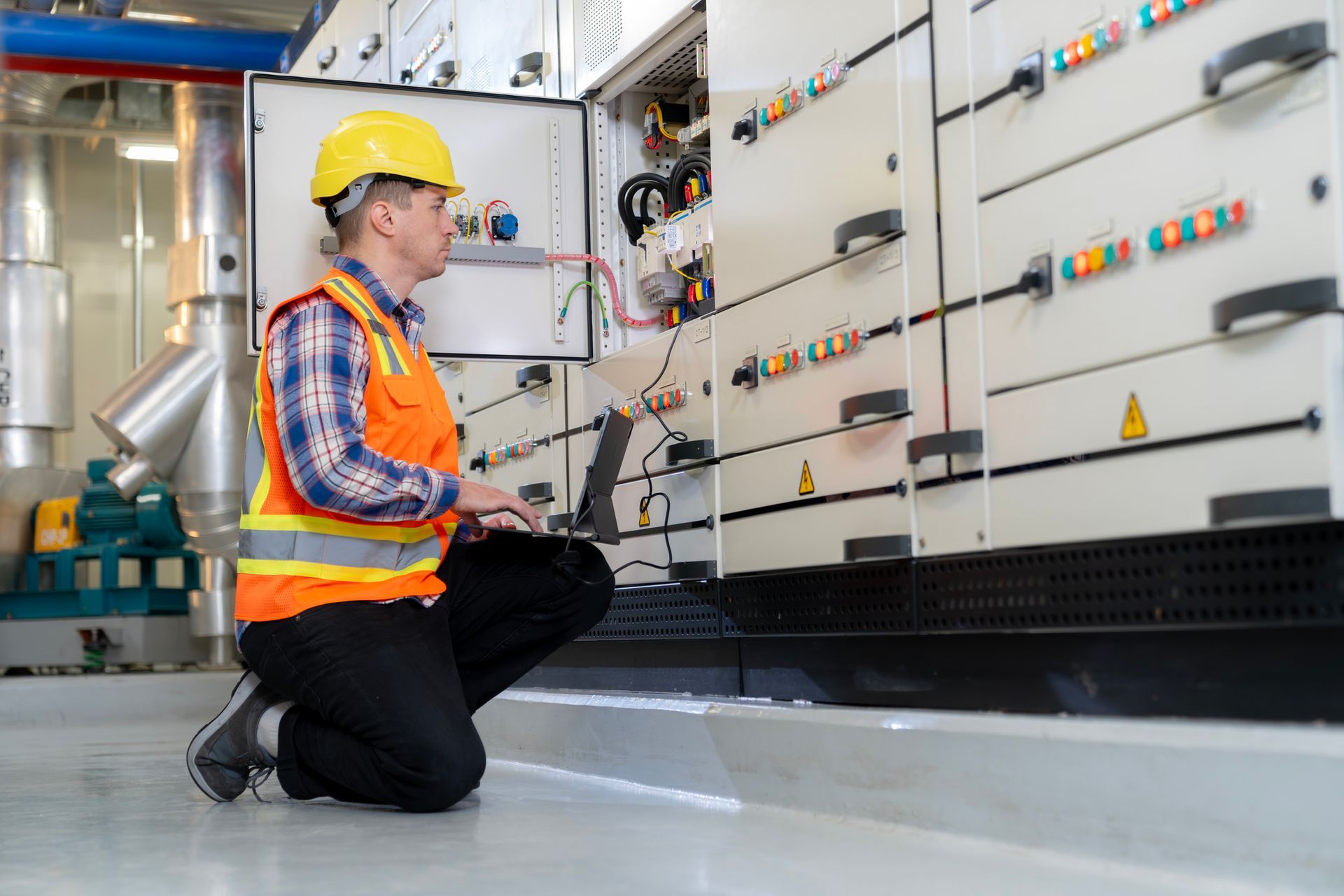 Electrician in yellow hard hat and orange vest inspects equipment panel with laptop.
