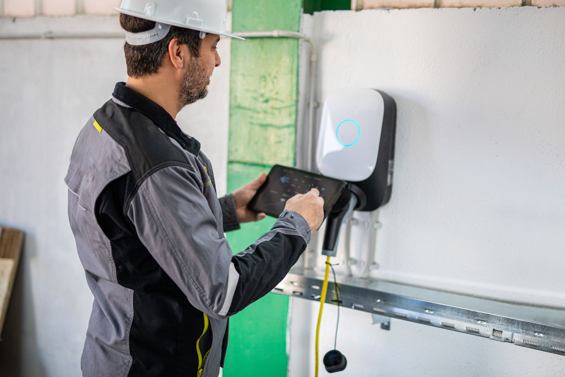 Construction worker in hard hat using a tablet to inspect an EV charger mounted on a wall.