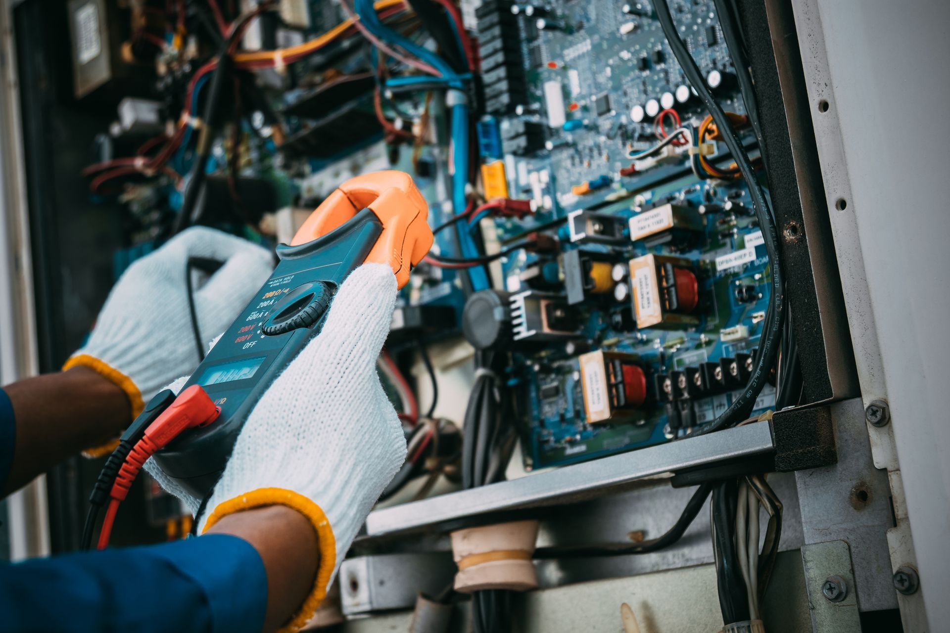 Person wearing gloves using a multimeter to inspect circuit board inside a machine.