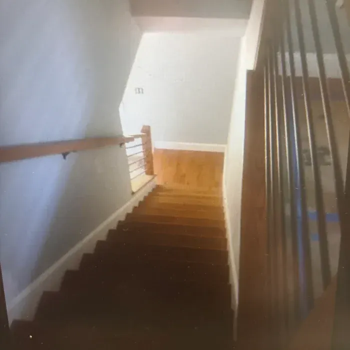 A wooden staircase looking down, with a gray wall on the left, white railings, and a landing at the bottom.