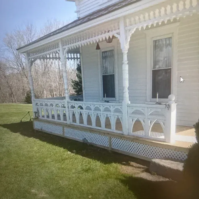 A white house with a detailed, arched wooden porch railing and decorative trim, set against a lawn and bare trees.