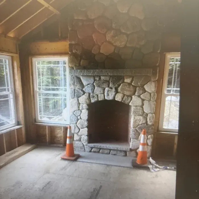A stone fireplace under construction in a room with unfinished wood walls and two windows, flanked by two traffic cones.