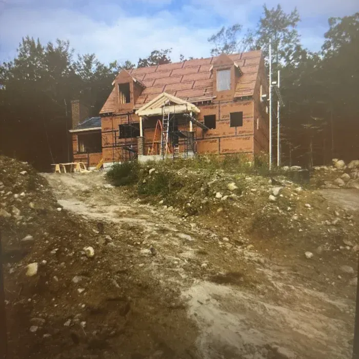 A house under construction with brown exterior sheathing, a gabled roof, dormers, and a rough, dirt-covered landscape.