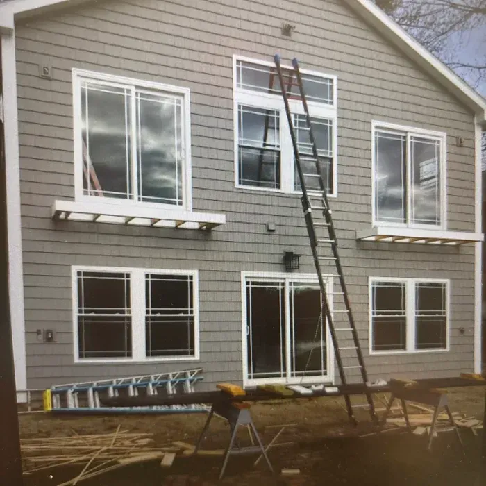 A two-story grey-sided house under construction with multiple windows, two ladders, and sawhorses in the yard.