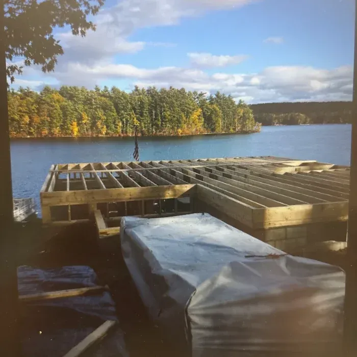 A wooden deck frame under construction overlooks a tranquil lake lined with autumn trees under a bright blue sky.