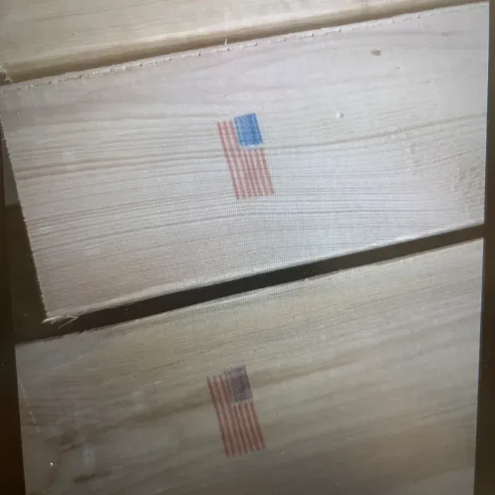 Two light-colored wooden boards stacked vertically, each stamped with a small, rectangular American flag icon.