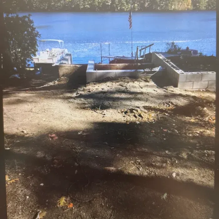 Construction of a concrete block foundation on a sandy slope overlooking a lake with a boat and dock.