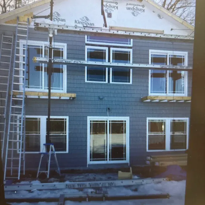 A two-story house under construction with grey siding, white-trimmed windows, a sliding glass door, and scaffolding.