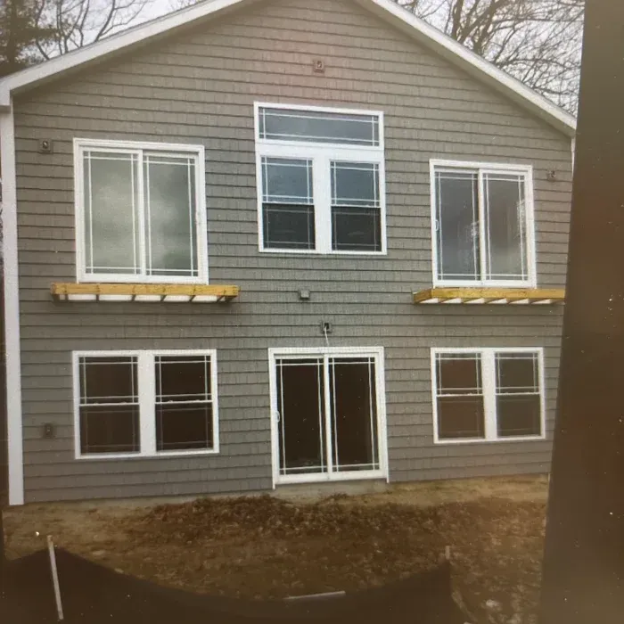 A two-story grey-sided house with a central glass door, surrounding windows, and unfinished wooden flower box supports.