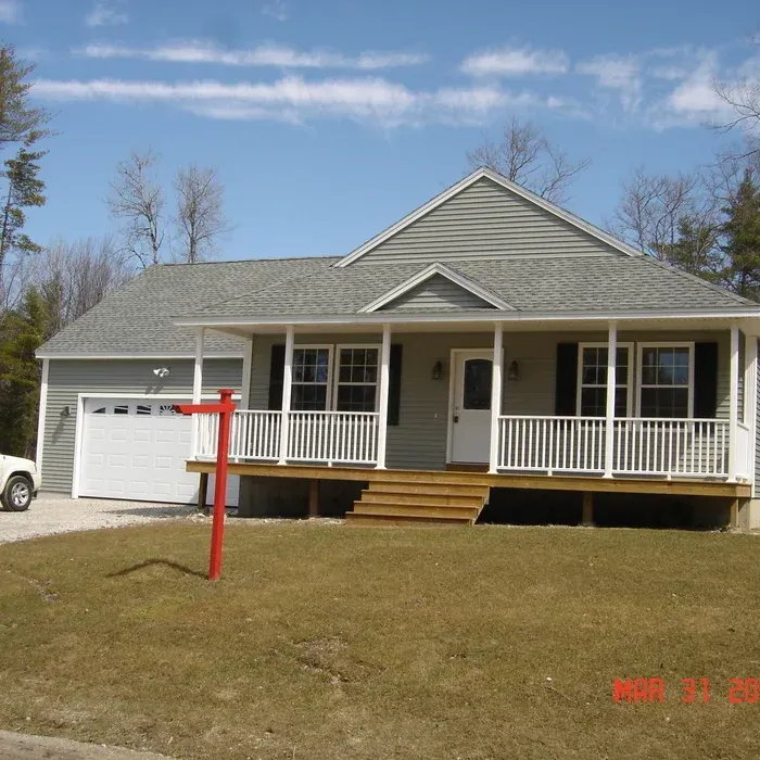 A light gray house with a front porch, white railings, and an attached garage under a blue sky, with a red real estate sign.