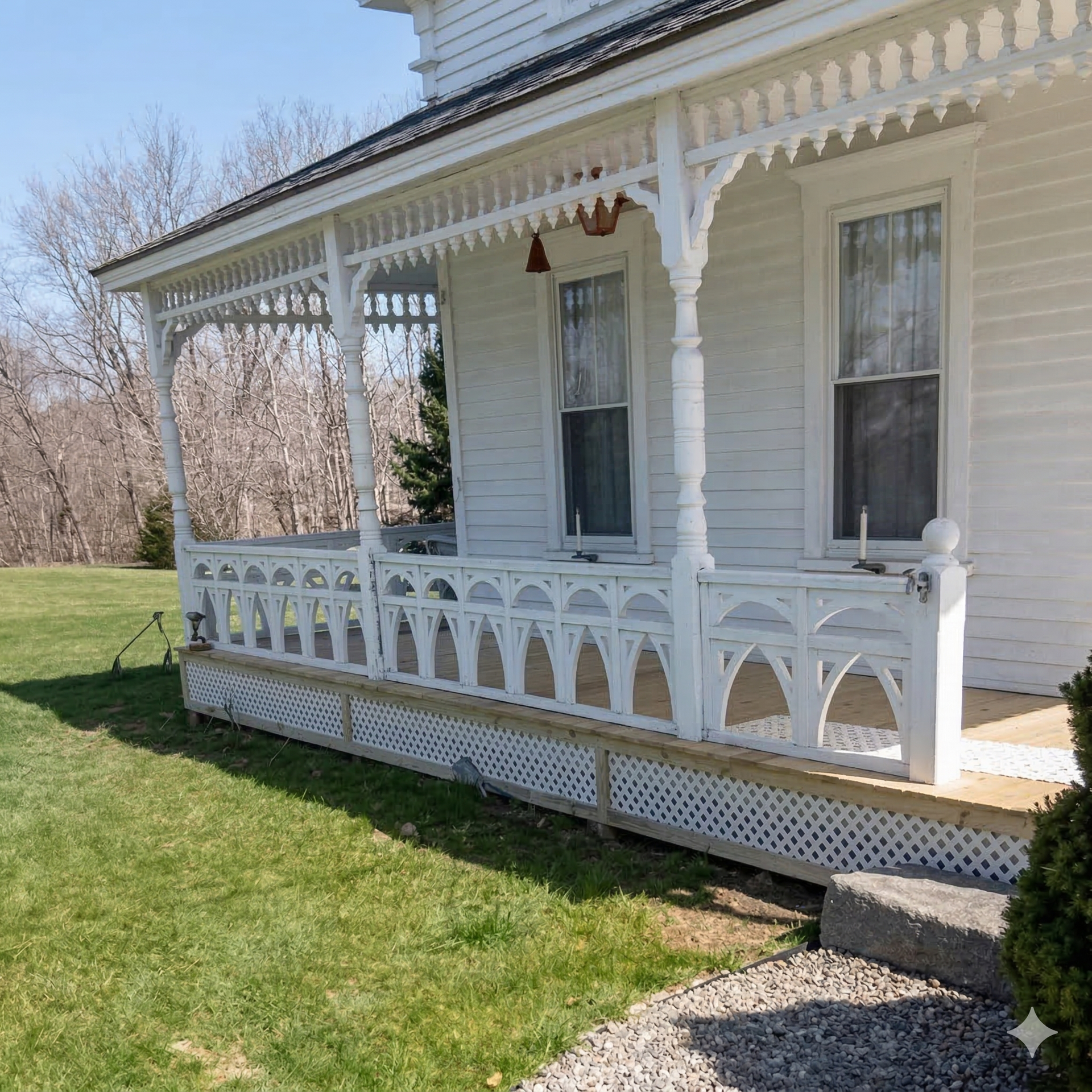 A white house with a porch featuring intricate, arched wooden railing and trim set against a lawn and bare trees.