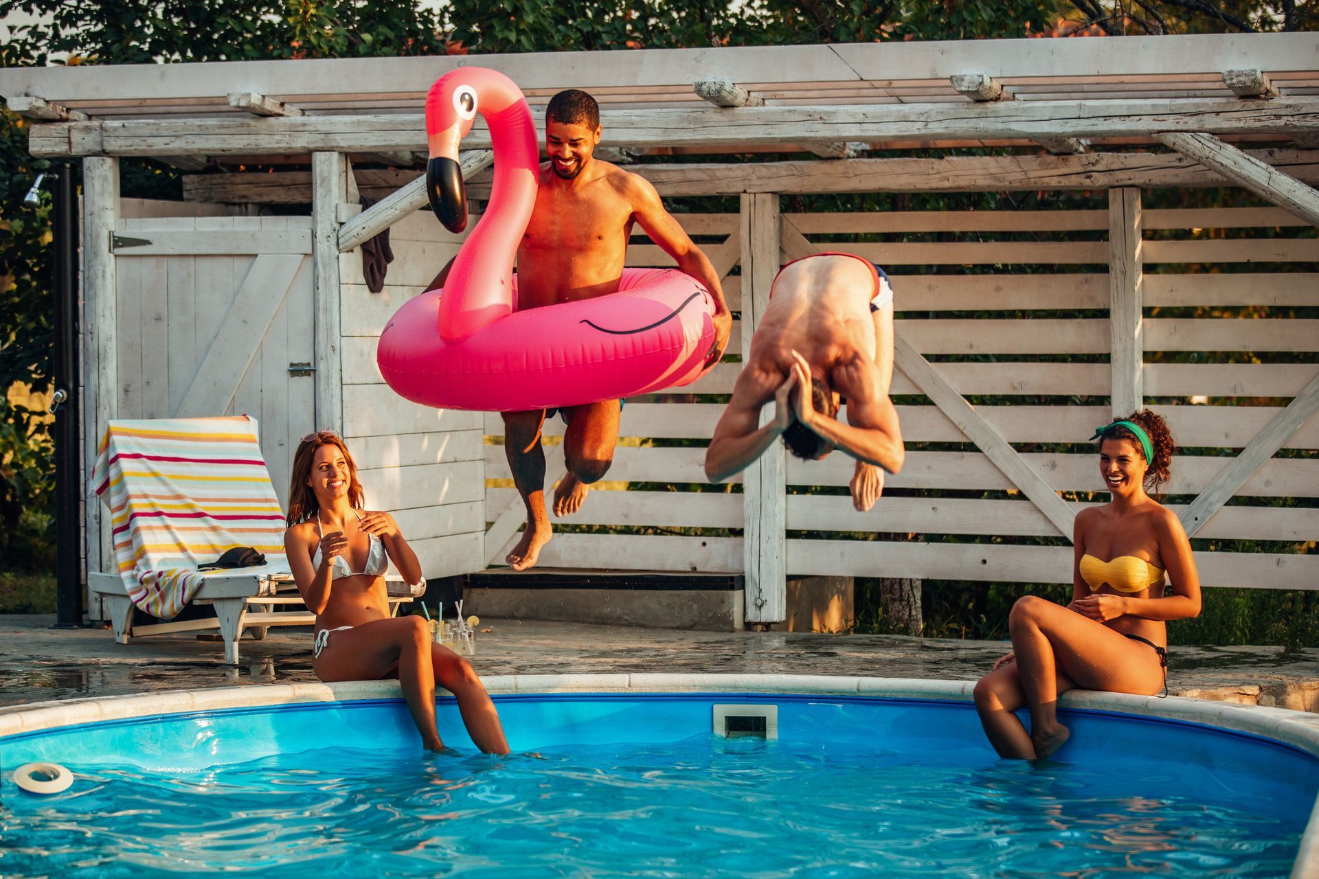 A group of people are jumping into a swimming pool with a flamingo float.