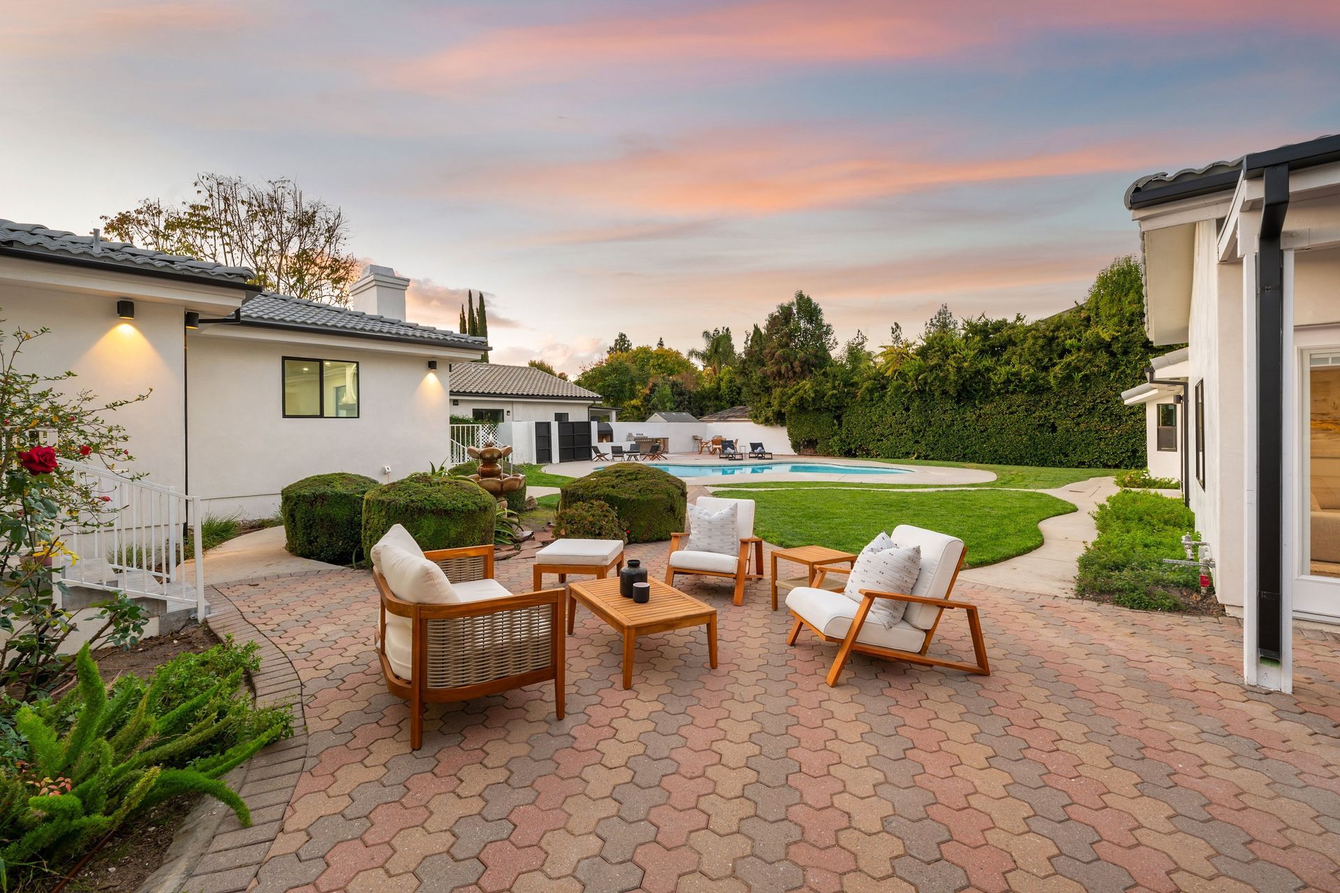 Patio with outdoor seating, brick paving, lush greenery, and pool under a colorful sky.
