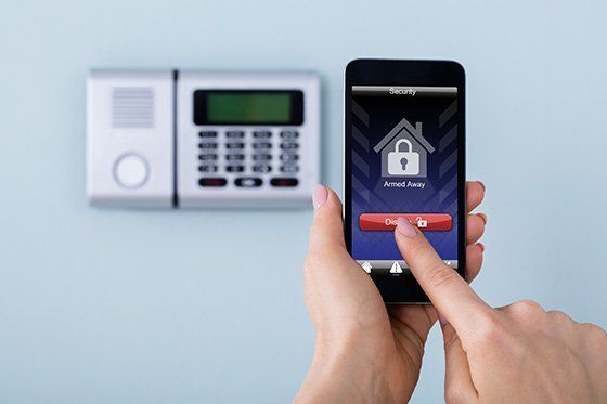 Persona che utilizza uno smartphone per controllare un sistema di sicurezza domestico; sullo sfondo si vede la tastiera.