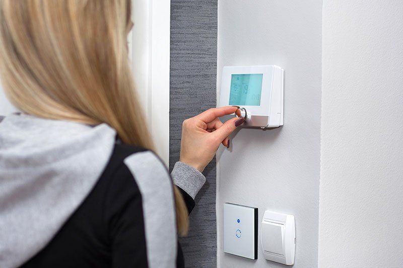 Donna che regola il termostato su una parete. Indossa una giacca con cappuccio ed è in piedi vicino al dispositivo.