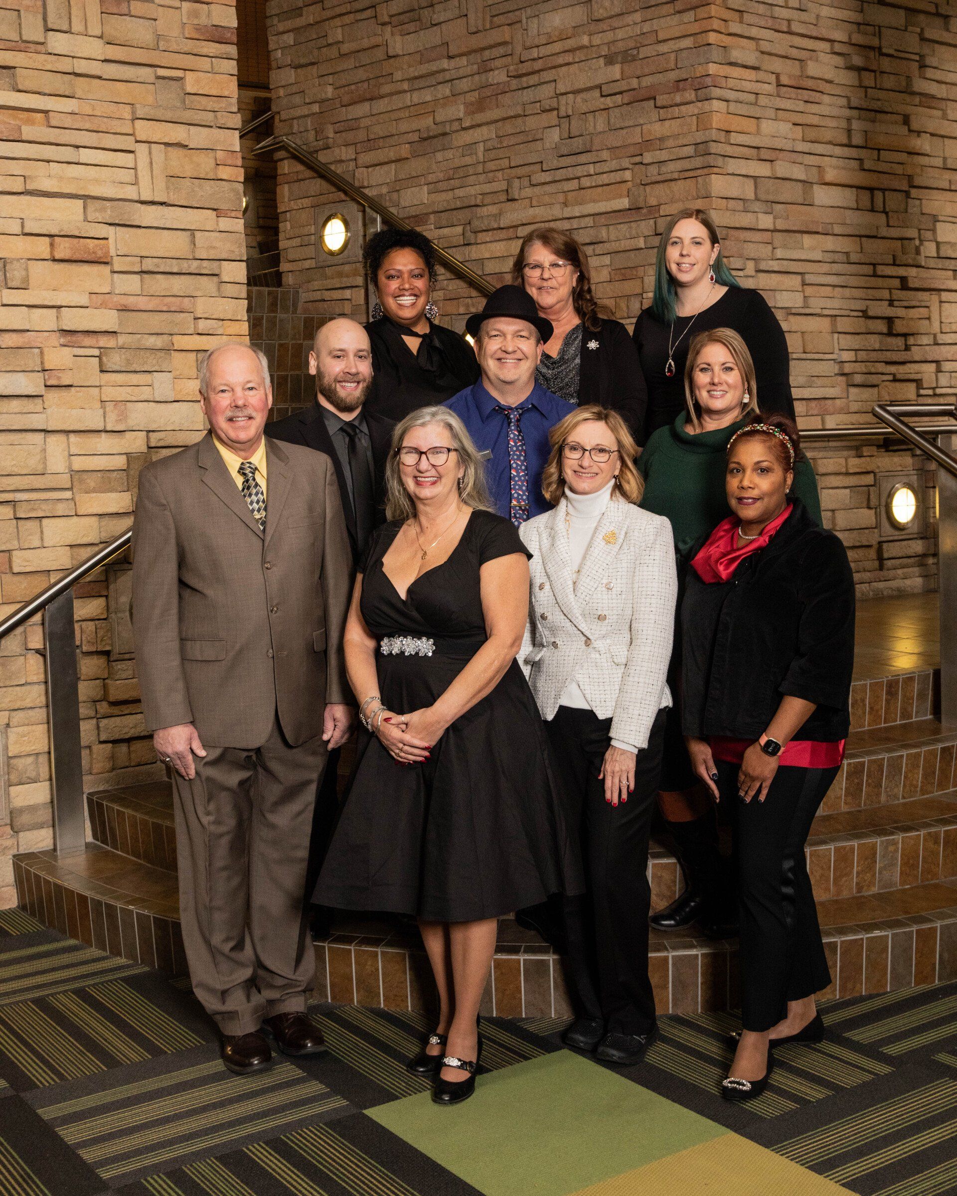 Group photo of diverse people posed on stairs, smiling. They are in formal attire in front of a textured wall.