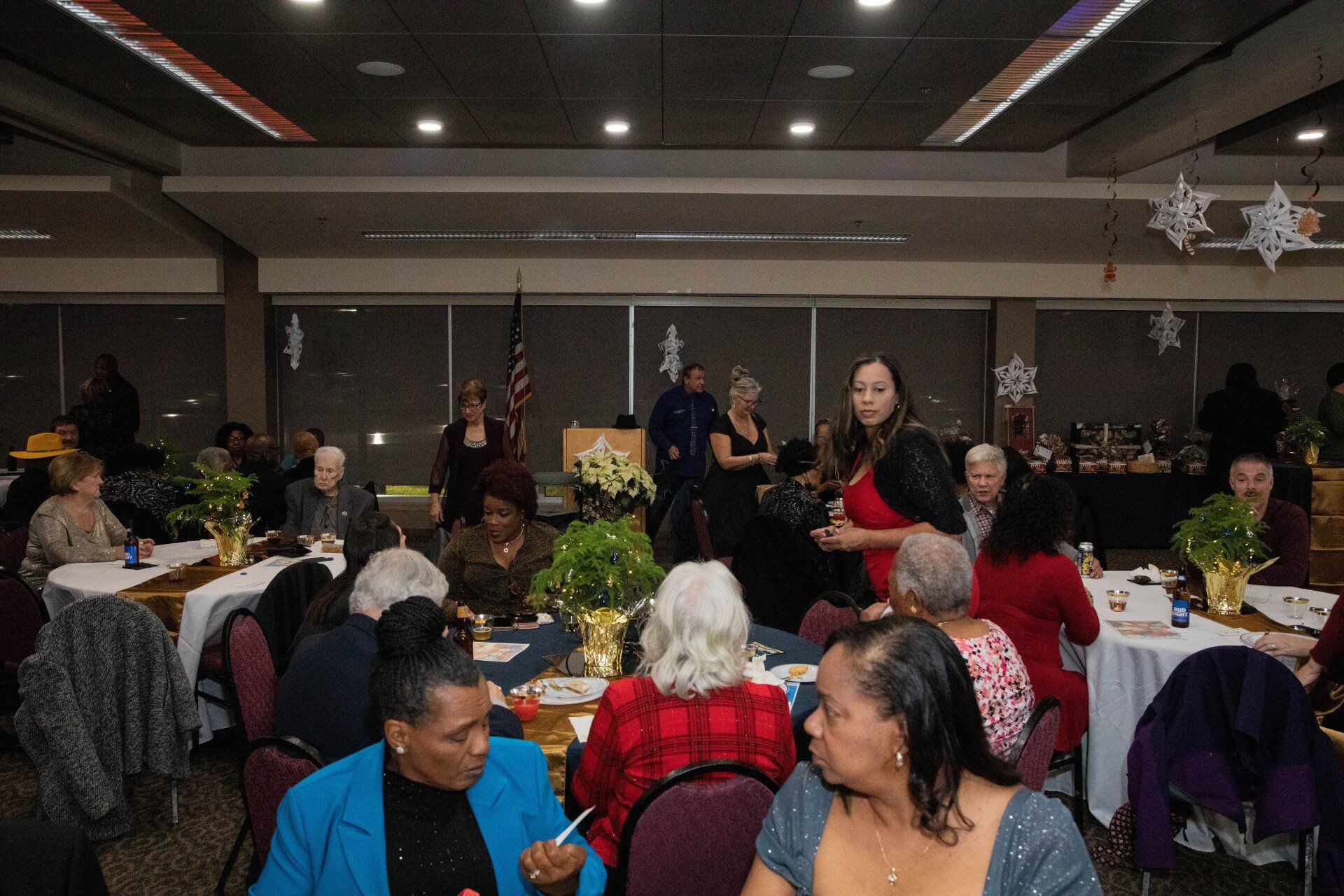 A festive banquet hall with seated guests at round tables, some wearing red. A woman in a red dress stands in the foreground.