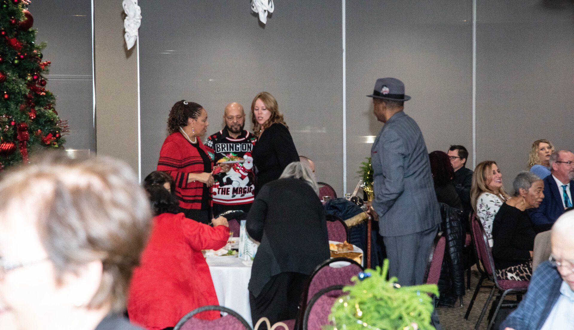 People at a holiday party; several are gathered around a table. A man in a fedora stands near the group, others are seated. A decorated Christmas tree is in the background.