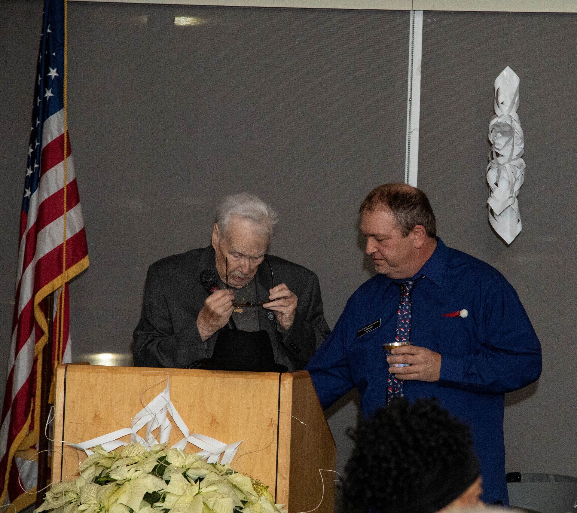 Two men at a podium with American flag and a white sculpture behind them. One man holds glasses, the other looks on.