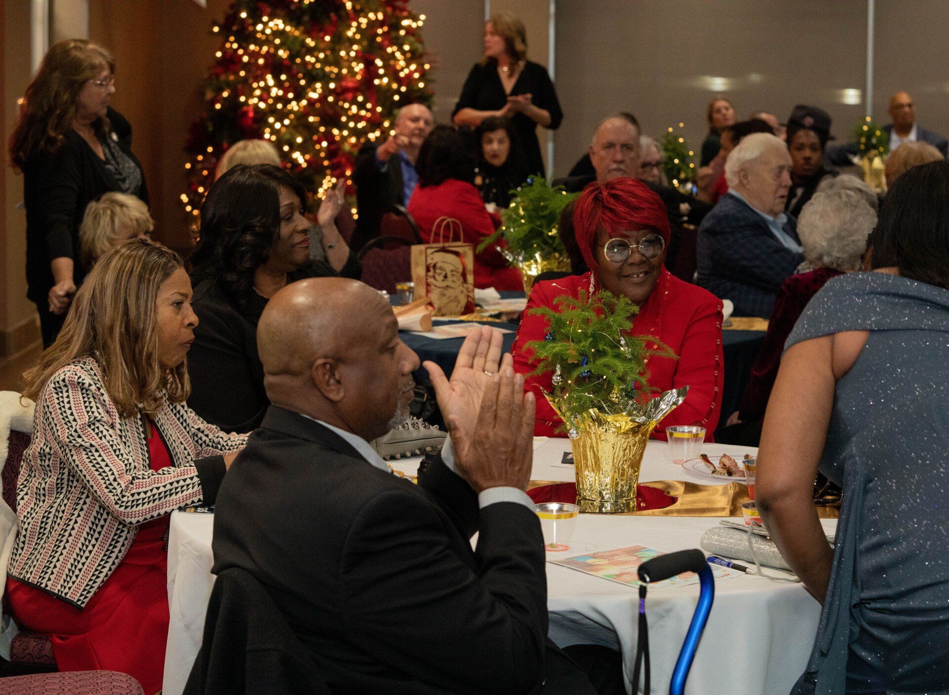 People at a festive gathering, clapping, smiling, and seated at tables decorated with centerpieces and in front of a Christmas tree.