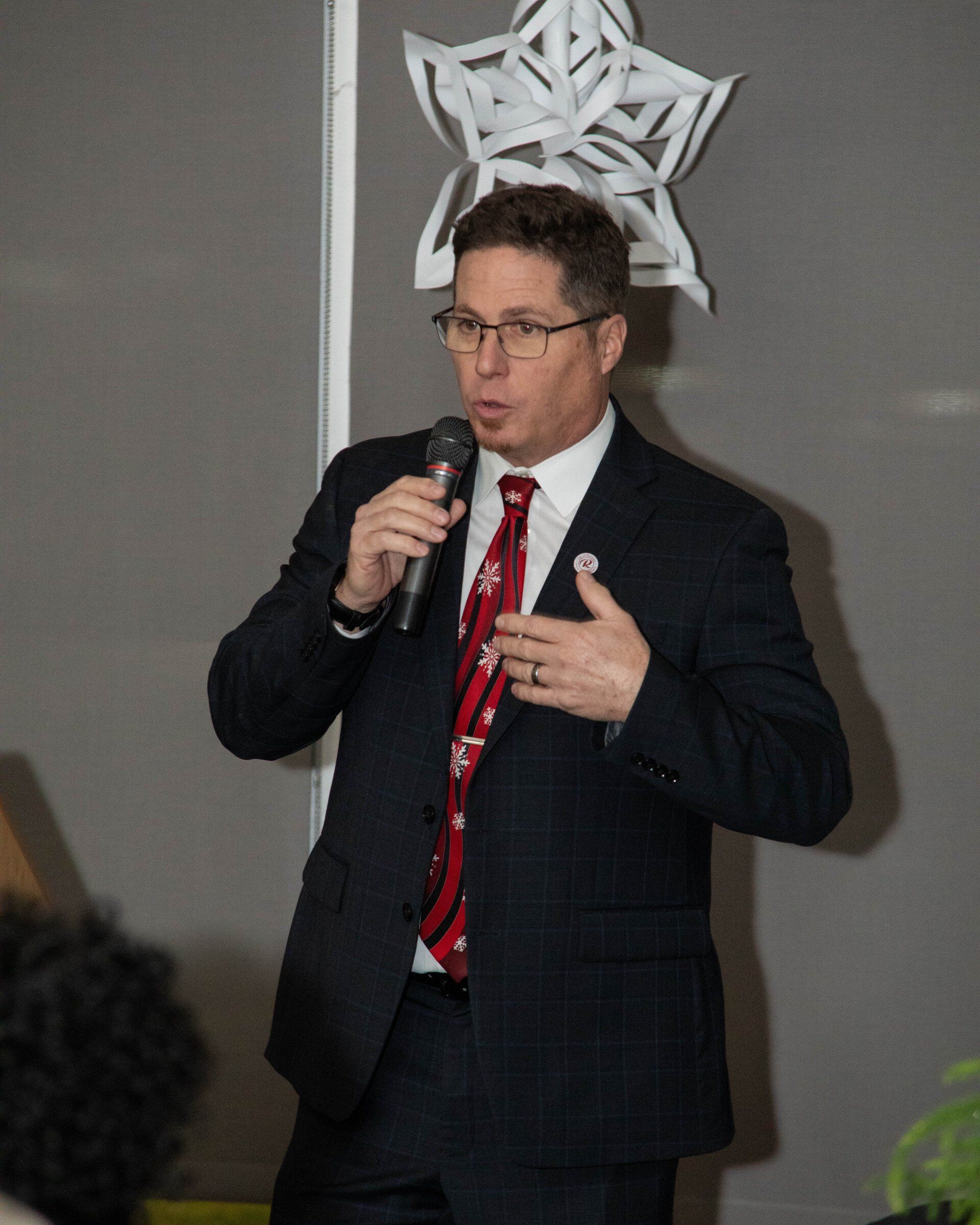 Man in suit speaks into microphone, gesturing with his right hand. Background with a star decoration.